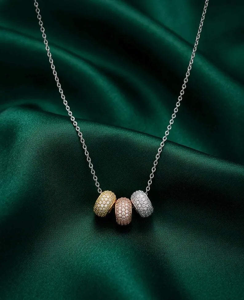 A delicate silver chain necklace with three small, textured capsule-shaped beads in yellow gold, rose gold, and silver, displayed on a draped dark green satin fabric.
