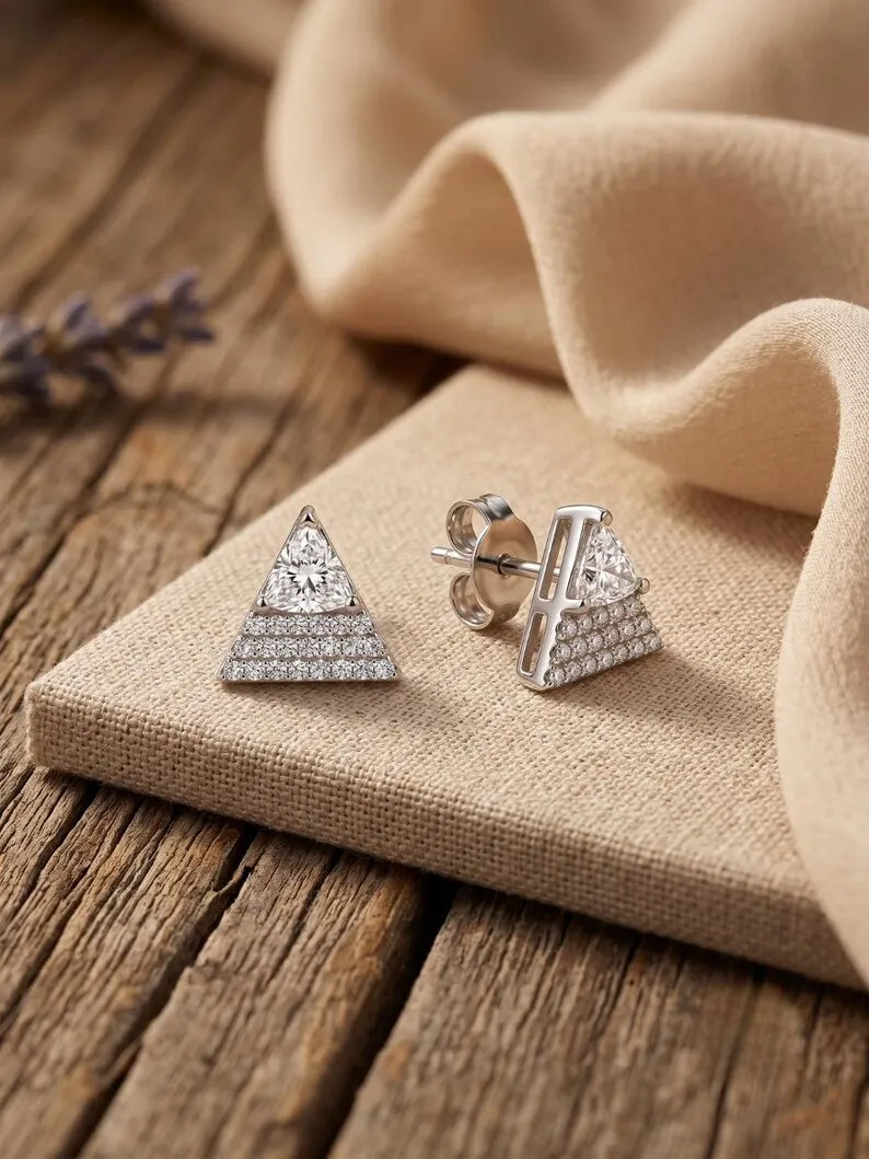 A pair of silver-toned triangle stud earrings with pavé-set clear stones, displayed on a textured beige fabric block over a rustic wooden surface.