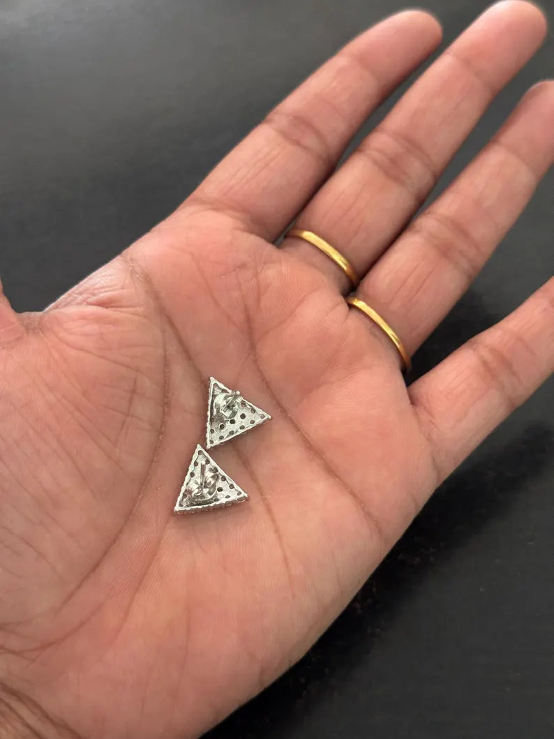 A close-up of a person's open palm holding a pair of small triangular silver stud earrings with clear stones, with two gold rings on the fingers against a dark background.