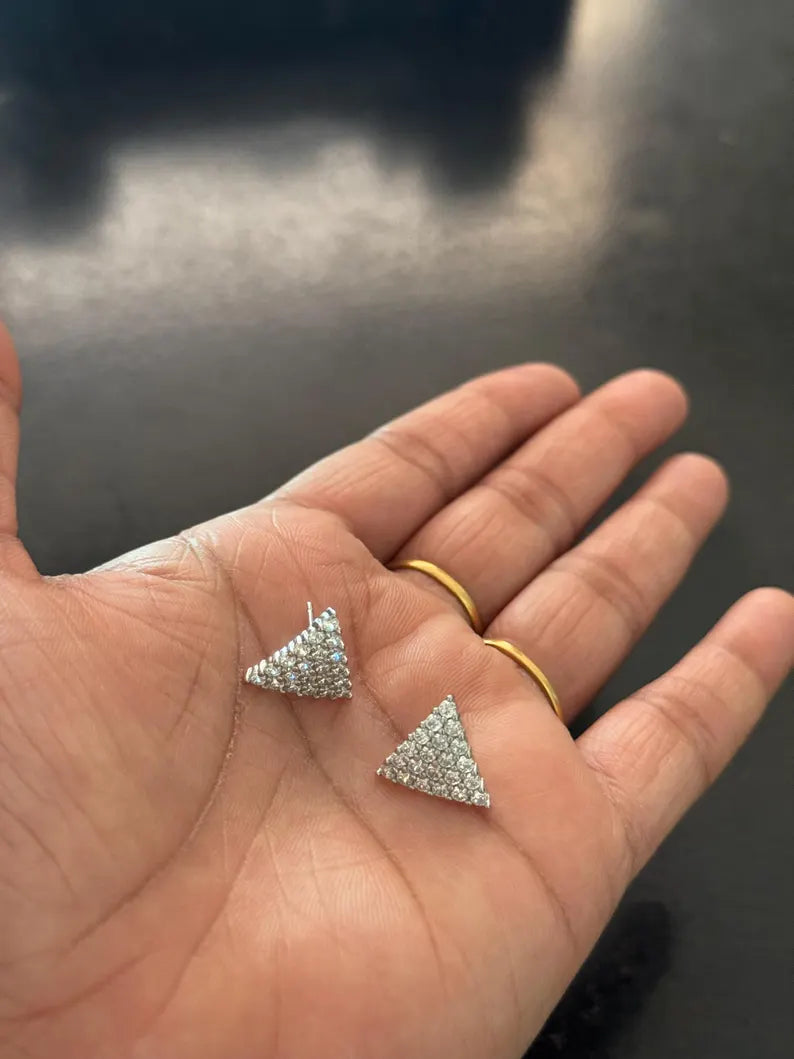 A close-up of a person's hand holding two small triangular stud earrings covered in clear stones, with two gold rings on the fingers against a dark blurred background.