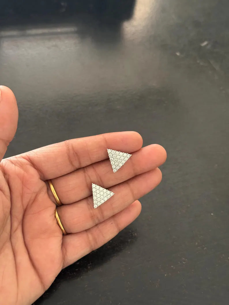 A close-up of a hand holding two small triangular stud earrings with a textured silver pattern, set against a dark gray background.