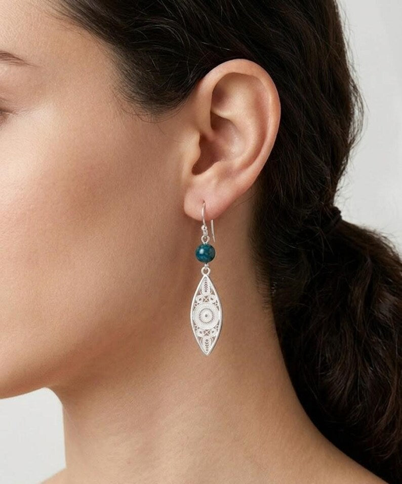 A close-up of a model's ear wearing a silver-toned dangle earring with a small dark teal bead and an eye-shaped pendant.