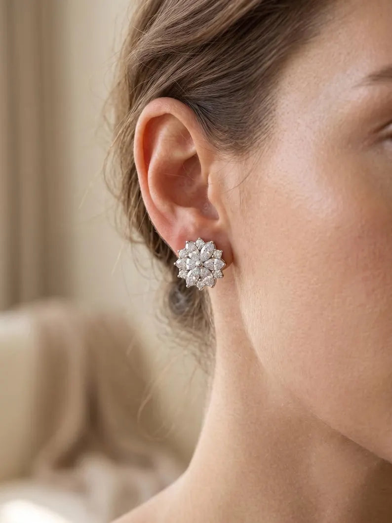 A close-up of a woman's ear wearing a silver-toned flower-shaped stud earring with clear gemstones, set against a softly blurred beige background.