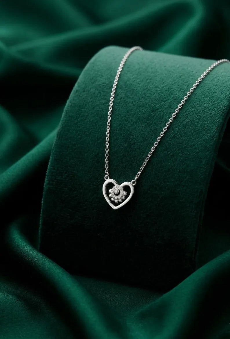 A silver heart-shaped pendant necklace with a small floral detail in the center, displayed on a dark green velvet jewelry stand.