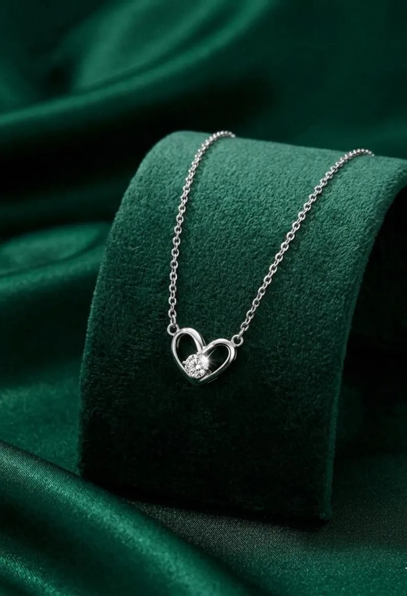 A silver heart pendant necklace with a small clear gemstone, displayed on a dark green velvet jewelry stand against a dark green satin fabric background.